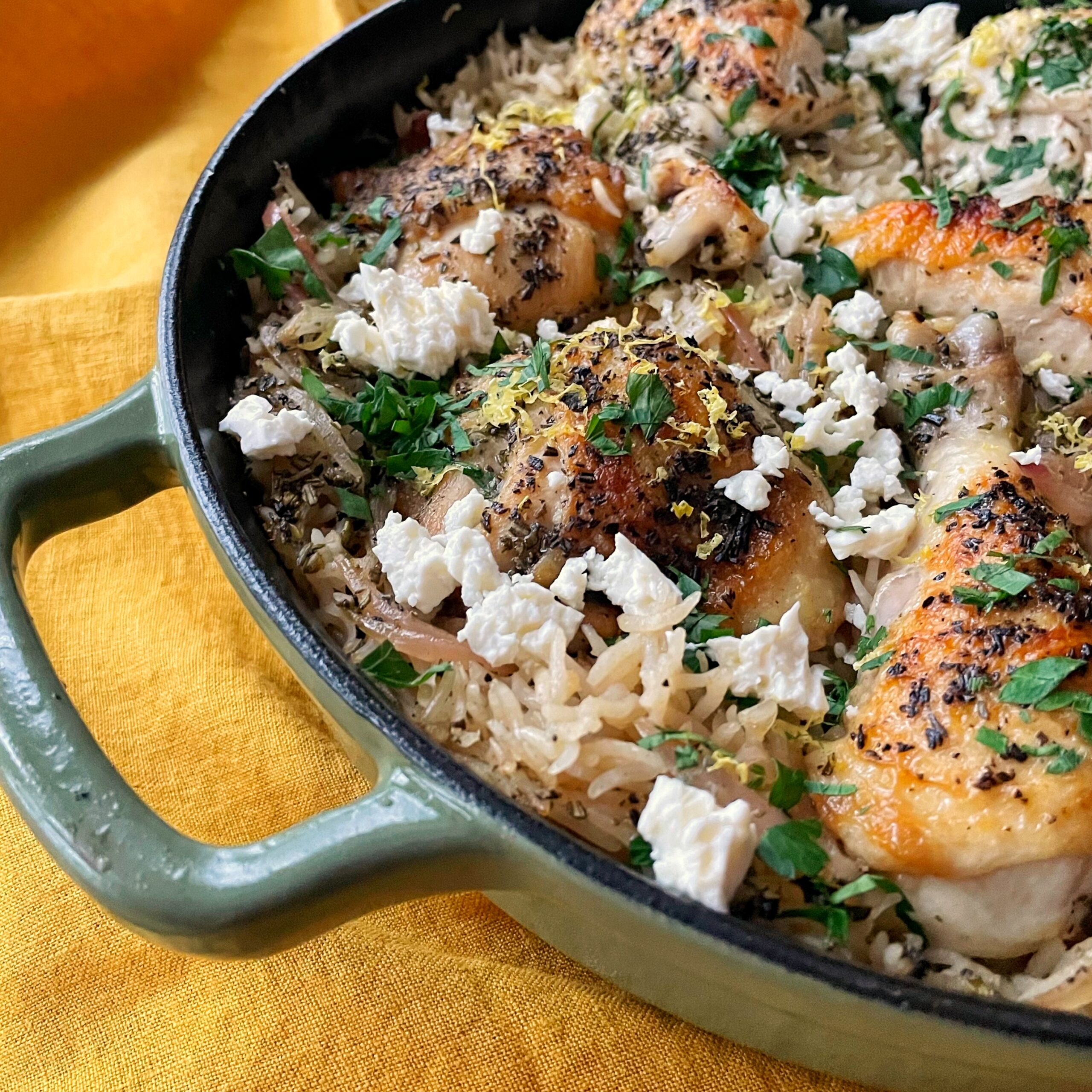 Green cast iron pan with chicken, rice, feta and parsley on a yellow backdrop.