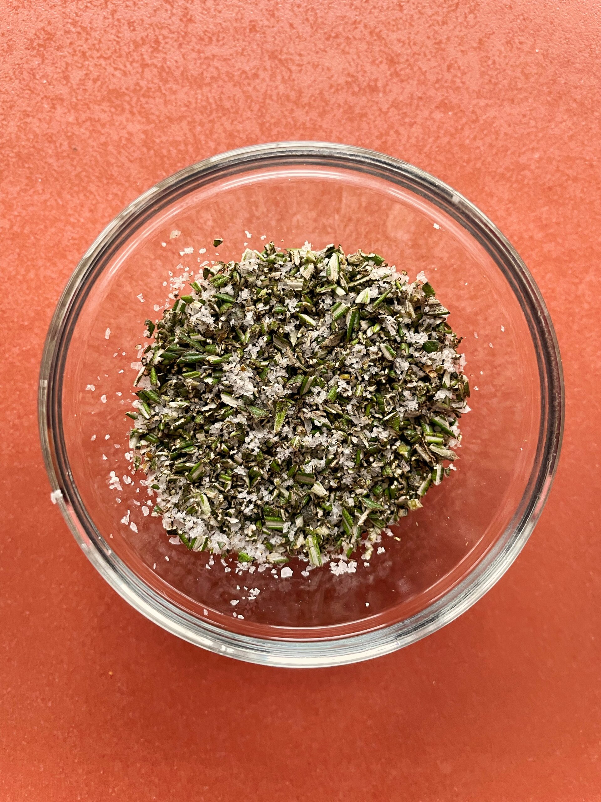 Finely chopped rosemary and salt in a glass dish on a pink backdrop.