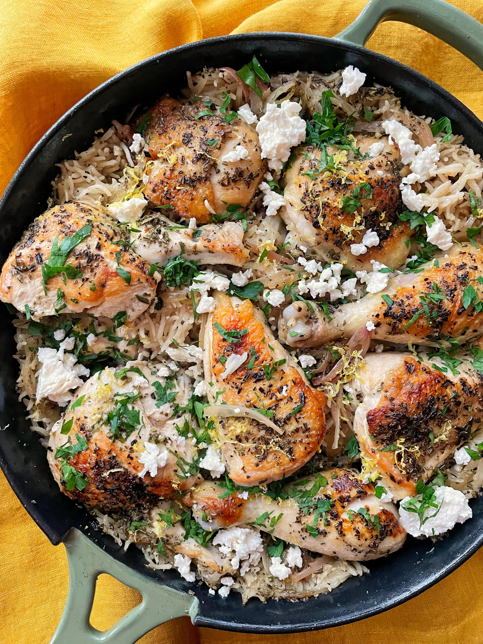 A green cast iron pan with rice, chicken feta and parsley sitting on a yellow cloth napkin.