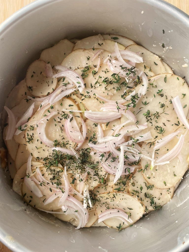 Cake pan with layers of potatoes topped with thinly sliced shallots and finely chopped thyme.