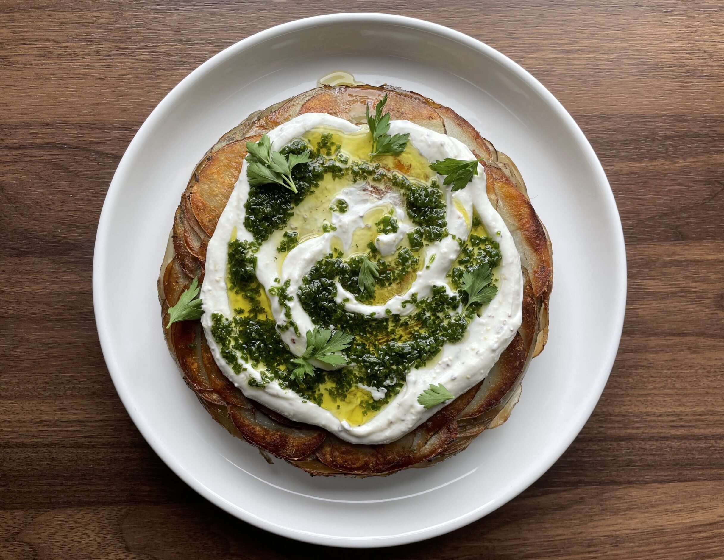 Circular potato cake with white and green sauce sitting on a white plate with a dark wooden background.