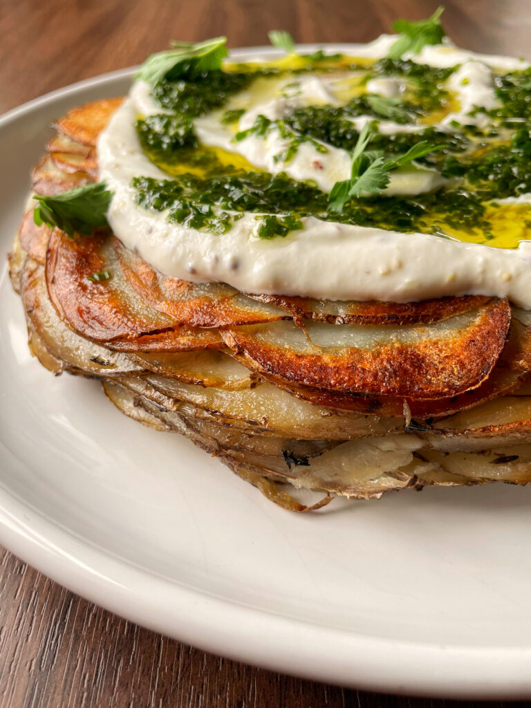 Fried potato cake topped with white yogurt sauce, green chive vinaigrette and fresh parsley on a white plate with a wooden background.