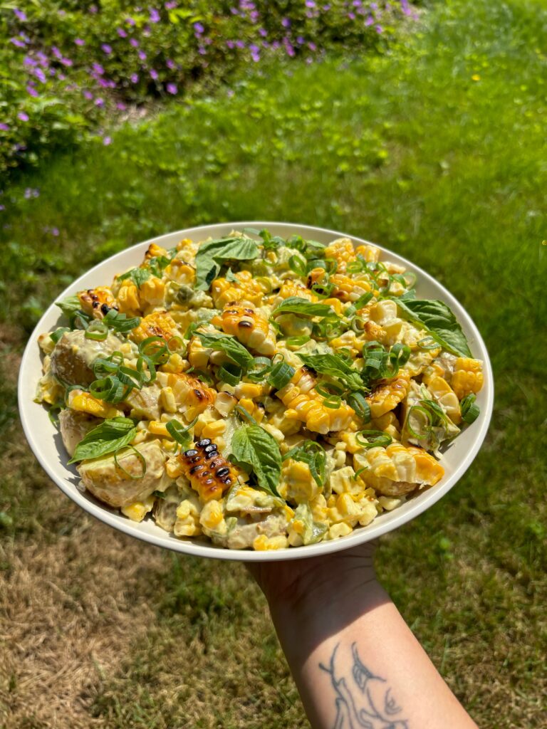 Hand holding serving plate of potato and charred corn salad outside with a grassy background.