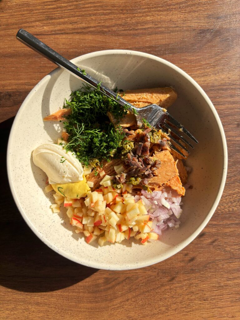 White bowl with black speckles holding tuna, mustard, mayo,diced apple, minched shallot, capers and freshly chopped dill.