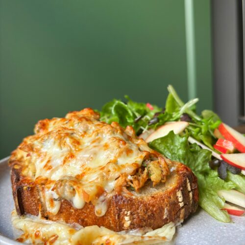 Bread with tuna salad and melted cheese beside a green salad on a large grey plate with a green background.