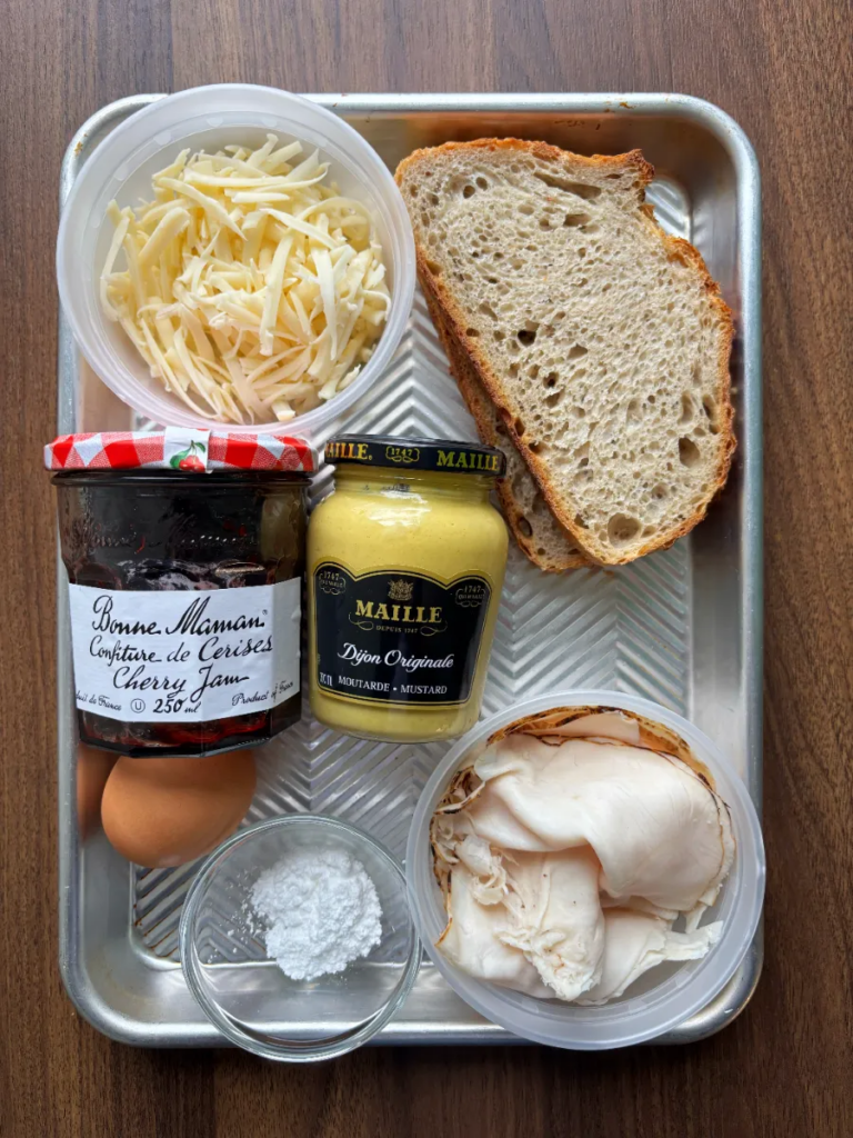 A metal sheet pan is sitting on a wooden table holding the ingredients for the Turkey and Cherry Monte Cristo. This includes cherry jam, dijon mustard, turkey, an egg, two slices of sour dough and icing sugar.