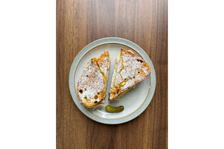 A light grey plate with a blue rim sitting on a wooden table holding a small pickle and a Monte Cristo Sandwich cut in half diagonally.