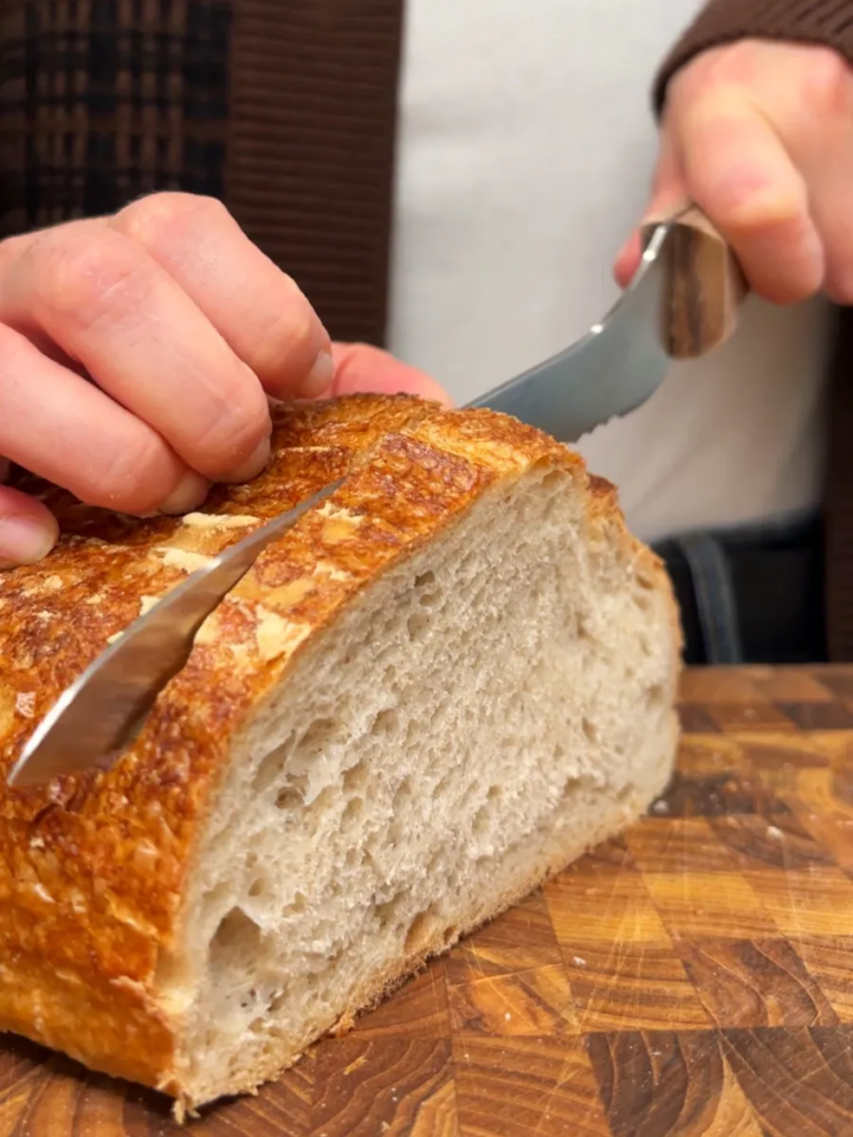 Sourdough bread being sliced with a serated knife on a fooden cutting board.