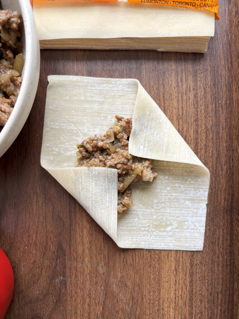 Egg roll wrapper on a wooden table topped with filling with the left and right edges fold in.