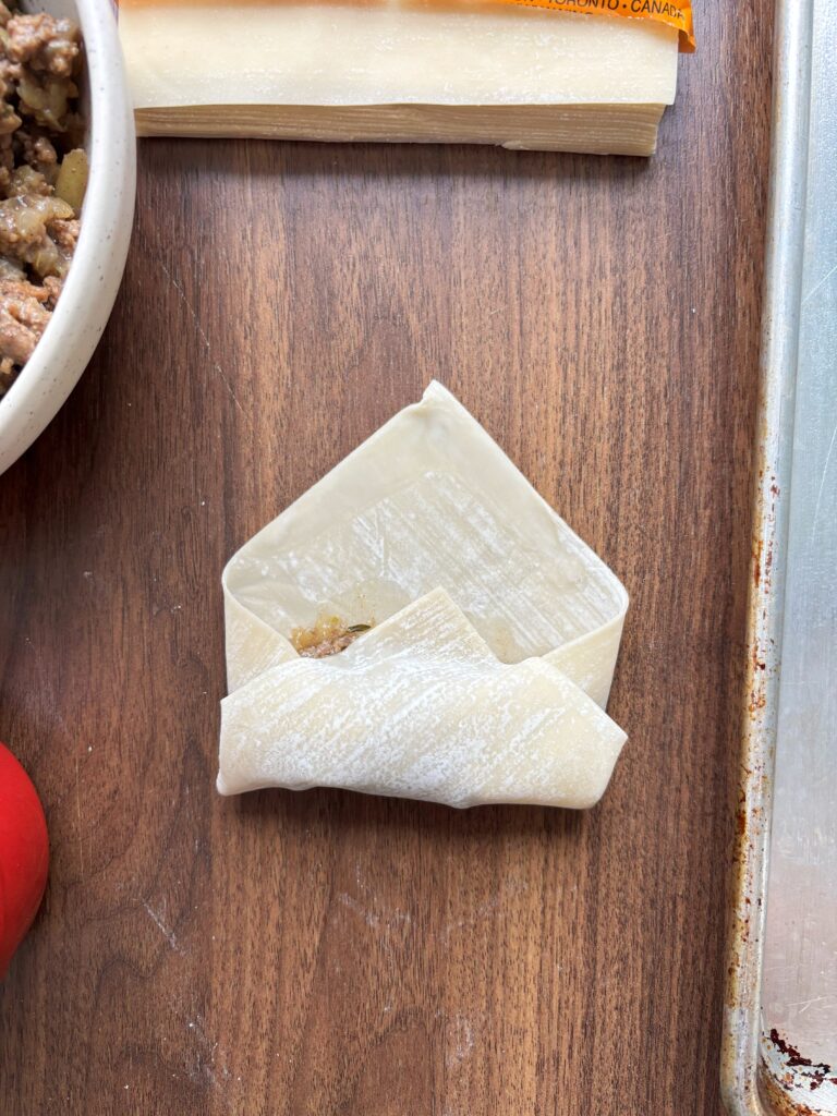 Tourtiere spring roll on a wooden table showing the step of tucking the wrapper under the filling.